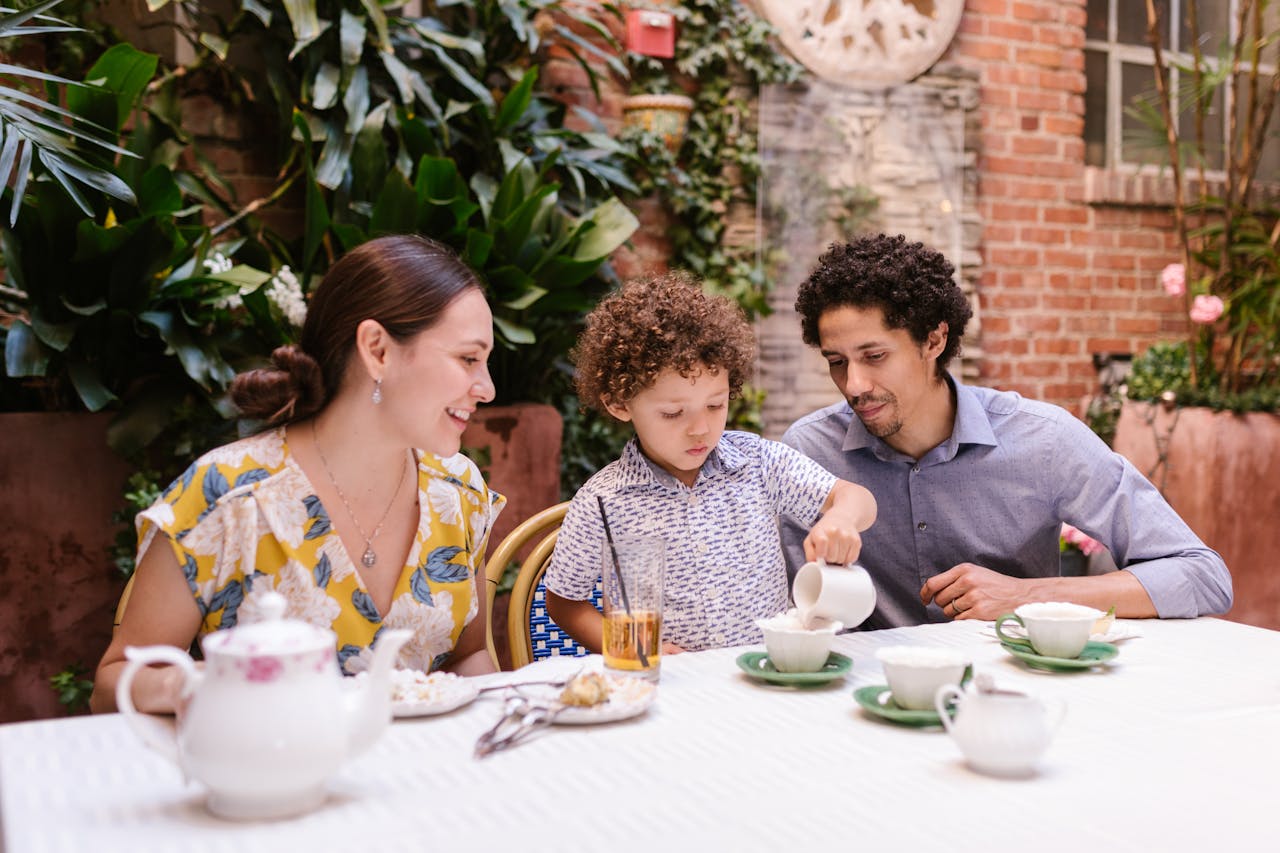 Smiling family enjoying tea time together at an outdoor café, with a young child pouring cream into a cup, symbolizing quality time and family connection.